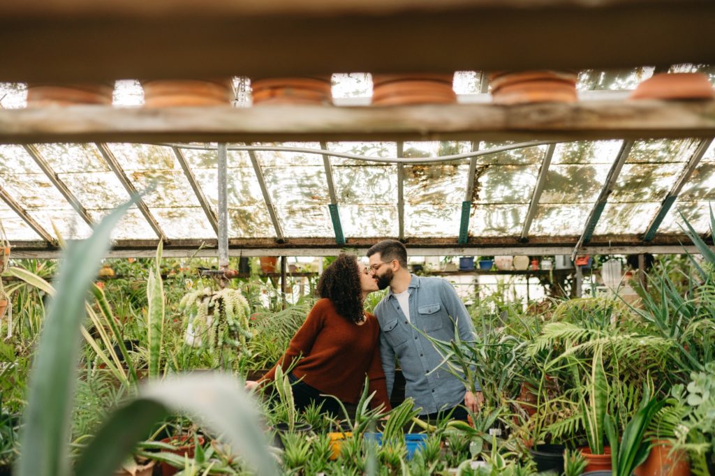 Clients kiss through hanging plants at John's Greenhouse Engagement Session